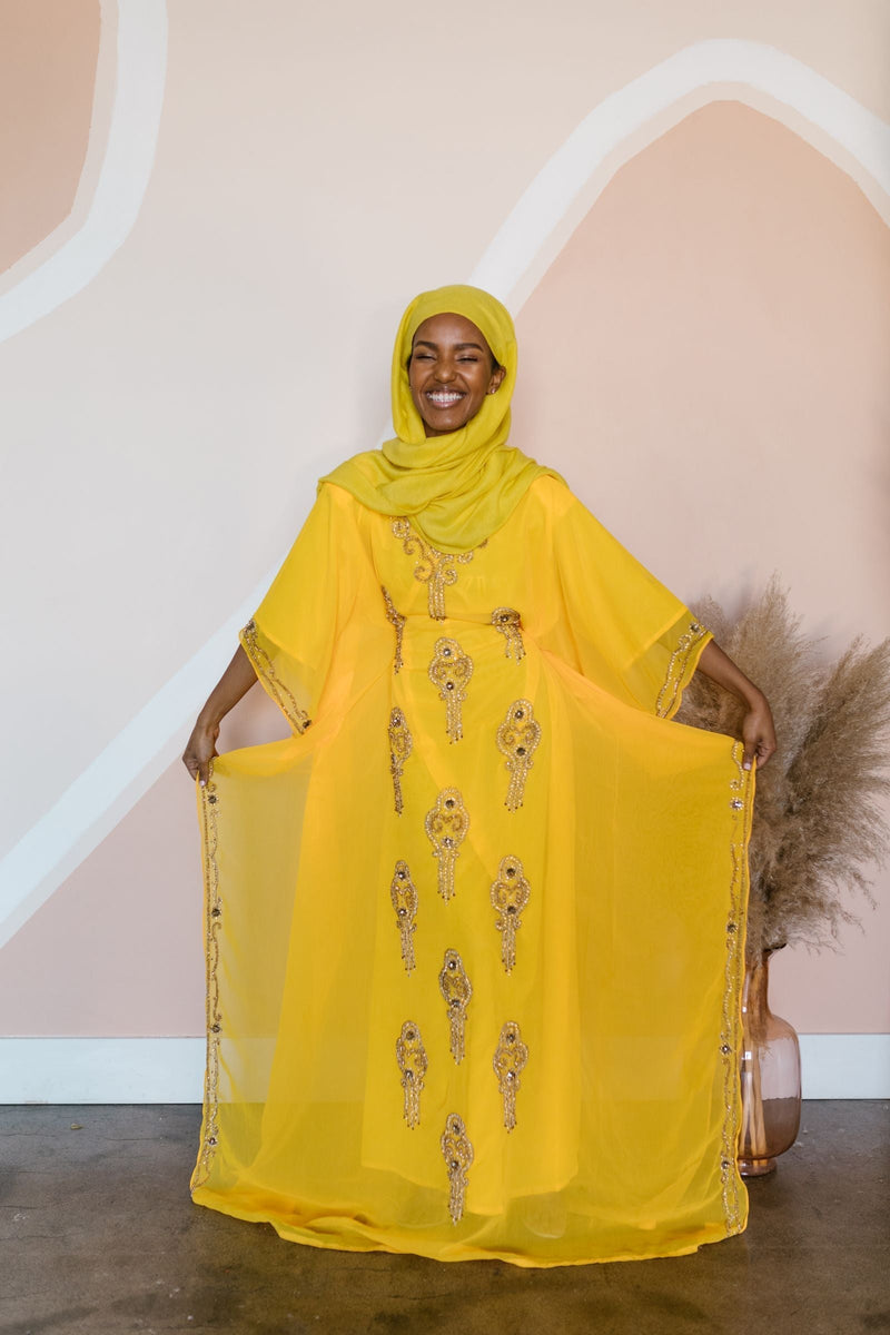 Smiling Muslim woman wearing a yellow long dress and matching hijab indoors, illustrating joyful modest Eid dress styling