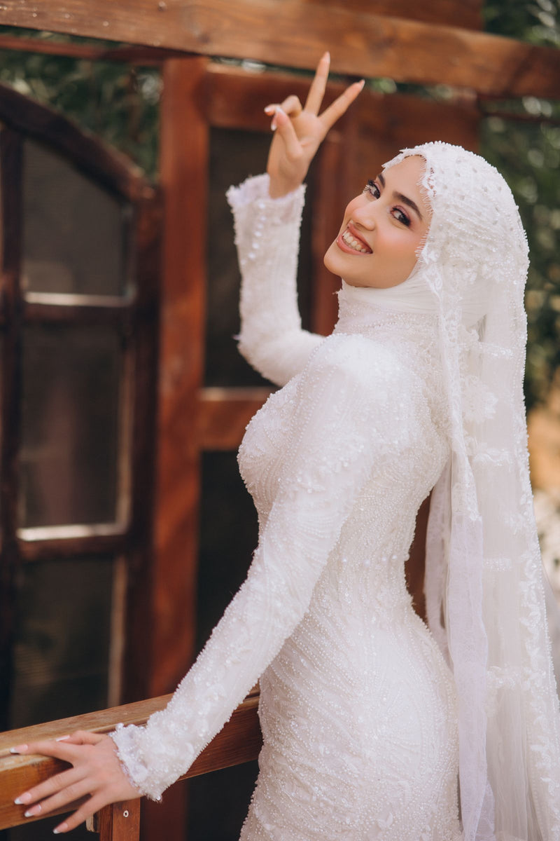 Muslim bride wearing a white long-sleeved wedding dress and hijab outdoors, illustrating modest bridal sleeves, neckline and gown fit
