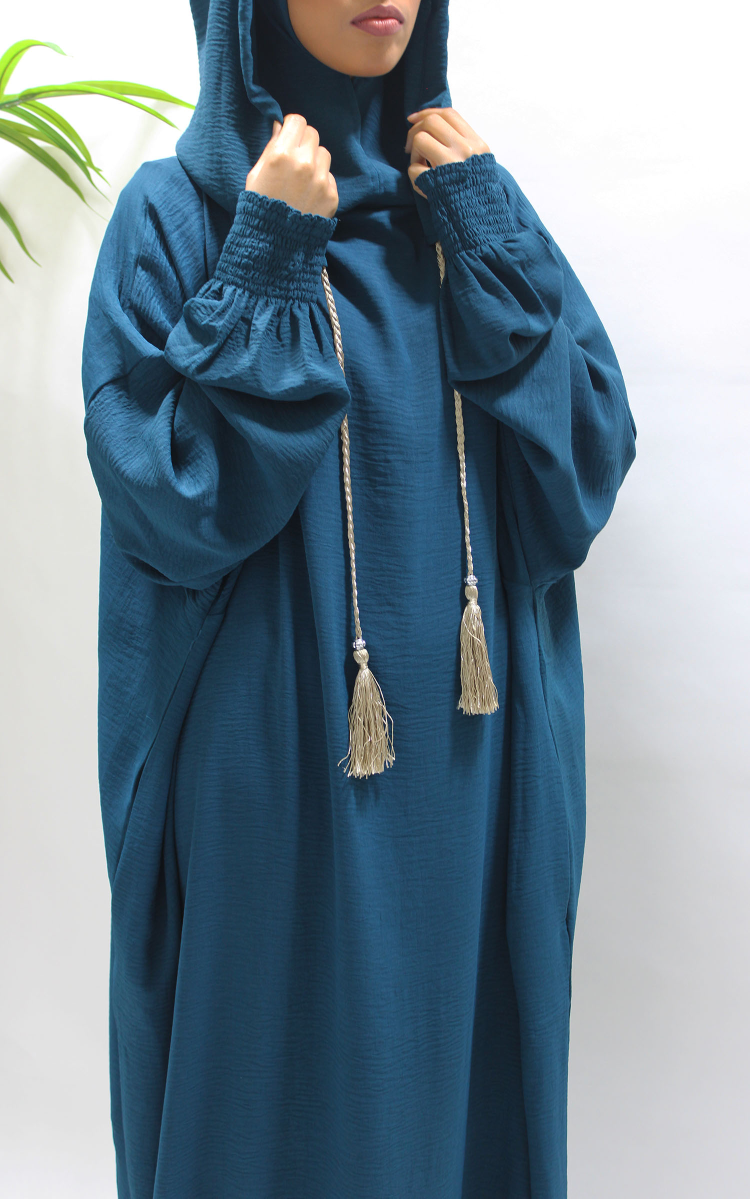  A modest woman wearing a teal double-hooded prayer abaya with ruched cuffs, standing confidently in a minimalist indoor setting, highlighting comfort and practicality.
